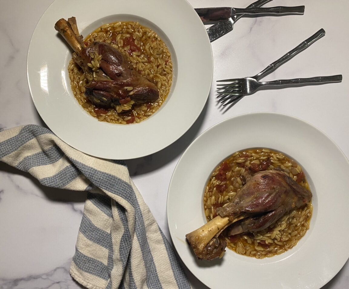 Lamb shank on top of orzo in a white bowl with a towel, forks, and knives