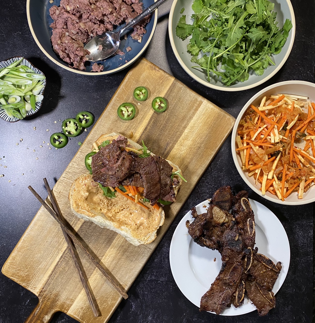 Asian short rib Banh mi sandwich on top of a cutting board Surround by a bowl of liver pate, arugula and cilantro, Banh mi slaw, and short ribs and jalapenos