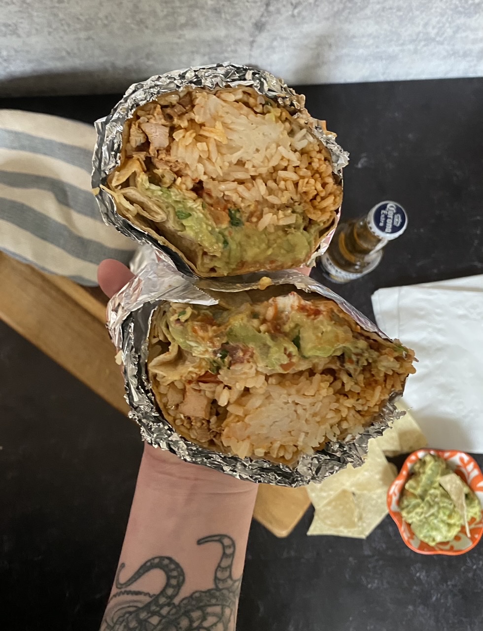 Two chicken burritos above a wood cutting board with a corona being held by a person with a tattoo