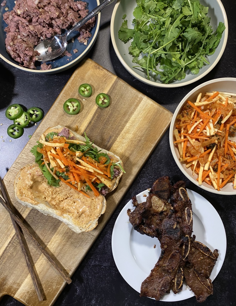 Asian short rib Banh mi sandwich on top of a cutting board Surround by a bowl of liver pate, arugula and cilantro, Banh mi slaw, and short ribs and jalapenos