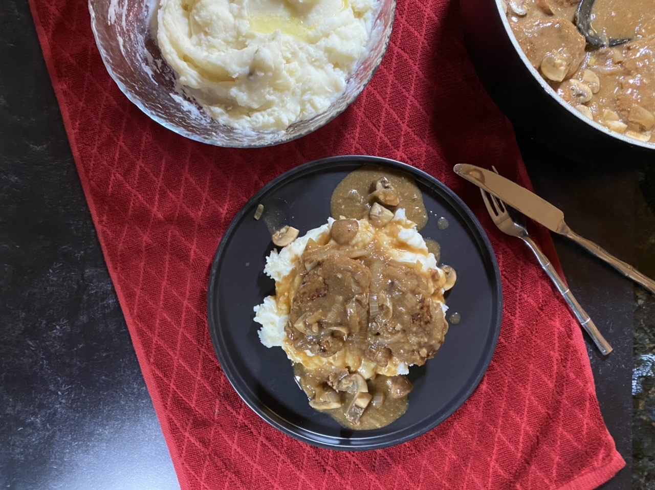Salisbury steak and gravy over mashed potatoes on a black plate on top of a red towel with a bowl of mashed potatoes and a sauce pan