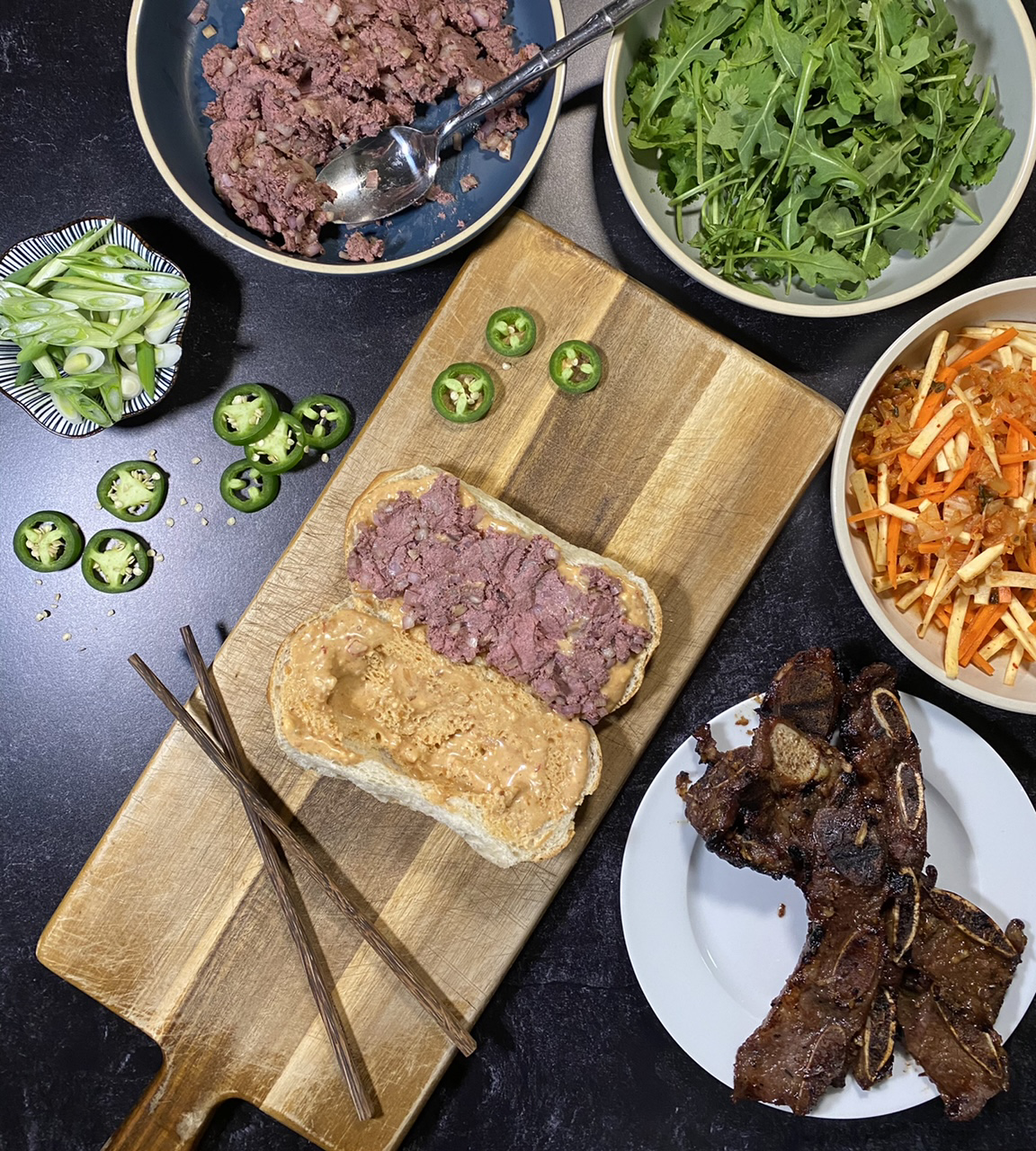 Asian short rib Banh mi sandwich on top of a cutting board Surround by a bowl of liver pate, arugula and cilantro, Banh mi slaw, and short ribs and jalapenos