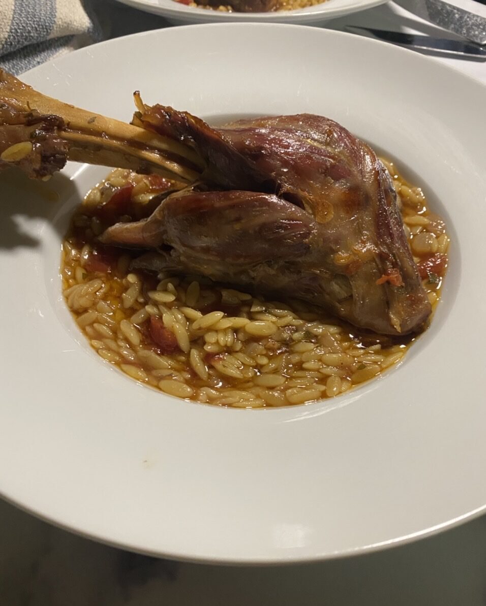 Lamb shank on top of orzo in a white bowl with a towel, forks, and knives