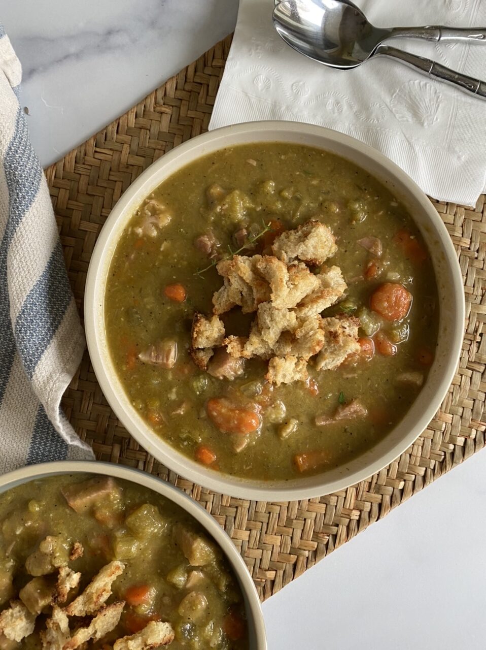 Two bowls of split pea soup with croutons and spoons on a woven mat