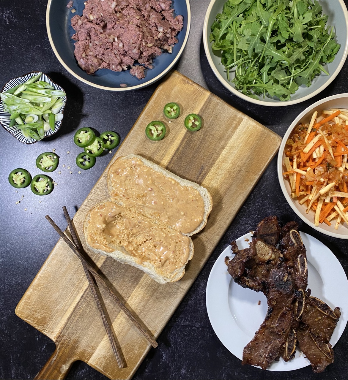 Asian short rib Banh mi sandwich on top of a cutting board Surround by a bowl of liver pate, arugula and cilantro, Banh mi slaw, and short ribs and jalapenos