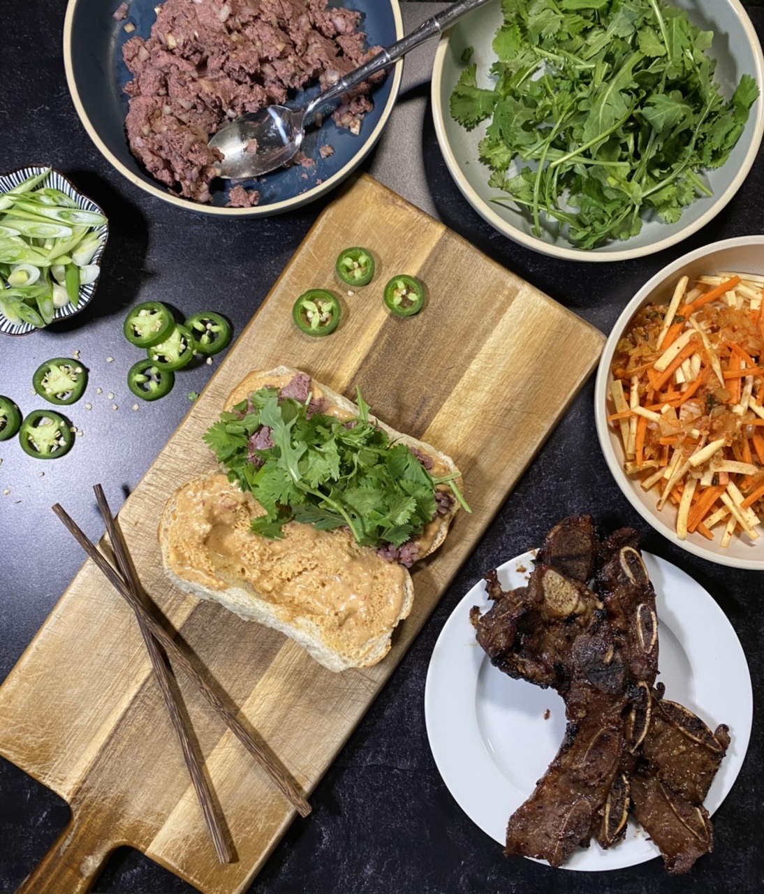 Asian short rib Banh mi sandwich on top of a cutting board Surround by a bowl of liver pate, arugula and cilantro, Banh mi slaw, and short ribs and jalapenos