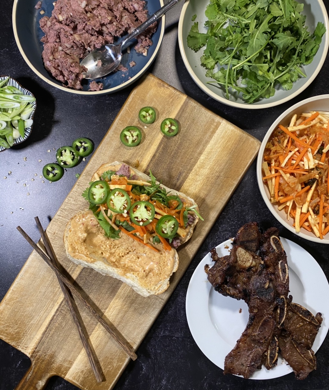 Asian short rib Banh mi sandwich on top of a cutting board Surround by a bowl of liver pate, arugula and cilantro, Banh mi slaw, and short ribs and jalapenos