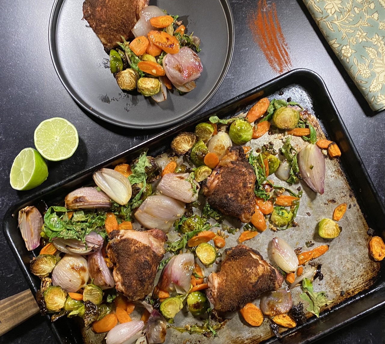 Chicken and vegetables on a baking sheet next to a black plate with chicken and vegetables