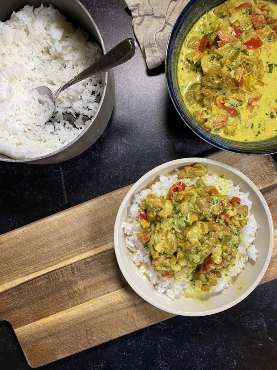 Bowl of Indian coconut curry chicken on top of rice next to a pot of rice and a bowl of coconut curry chicken