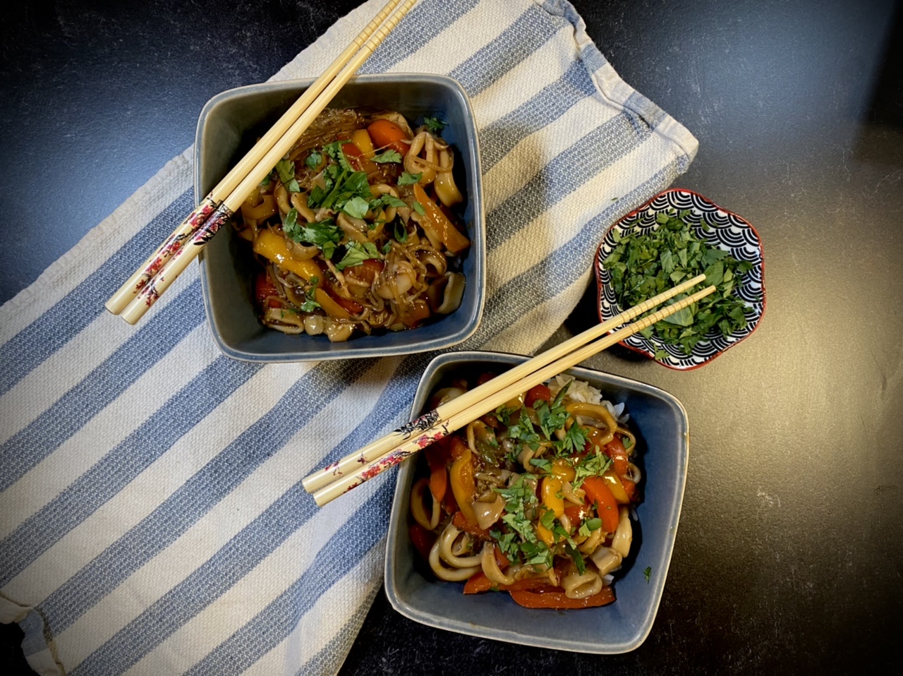 Squid stir fry in two blue bowls with chopsticks on top of a blue and white striped towel