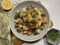 Chicken fricassee on a blue plate on top of a wooden cutting board next to a towel, lemon, and bowl of parsley