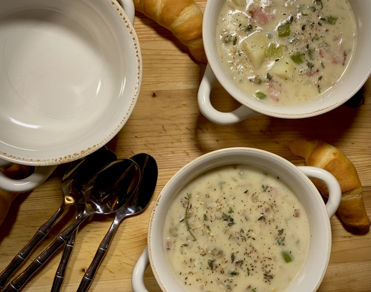 Two bowls of New England clam chowder on top of a wooden cutting board with spoons and crescent rolls