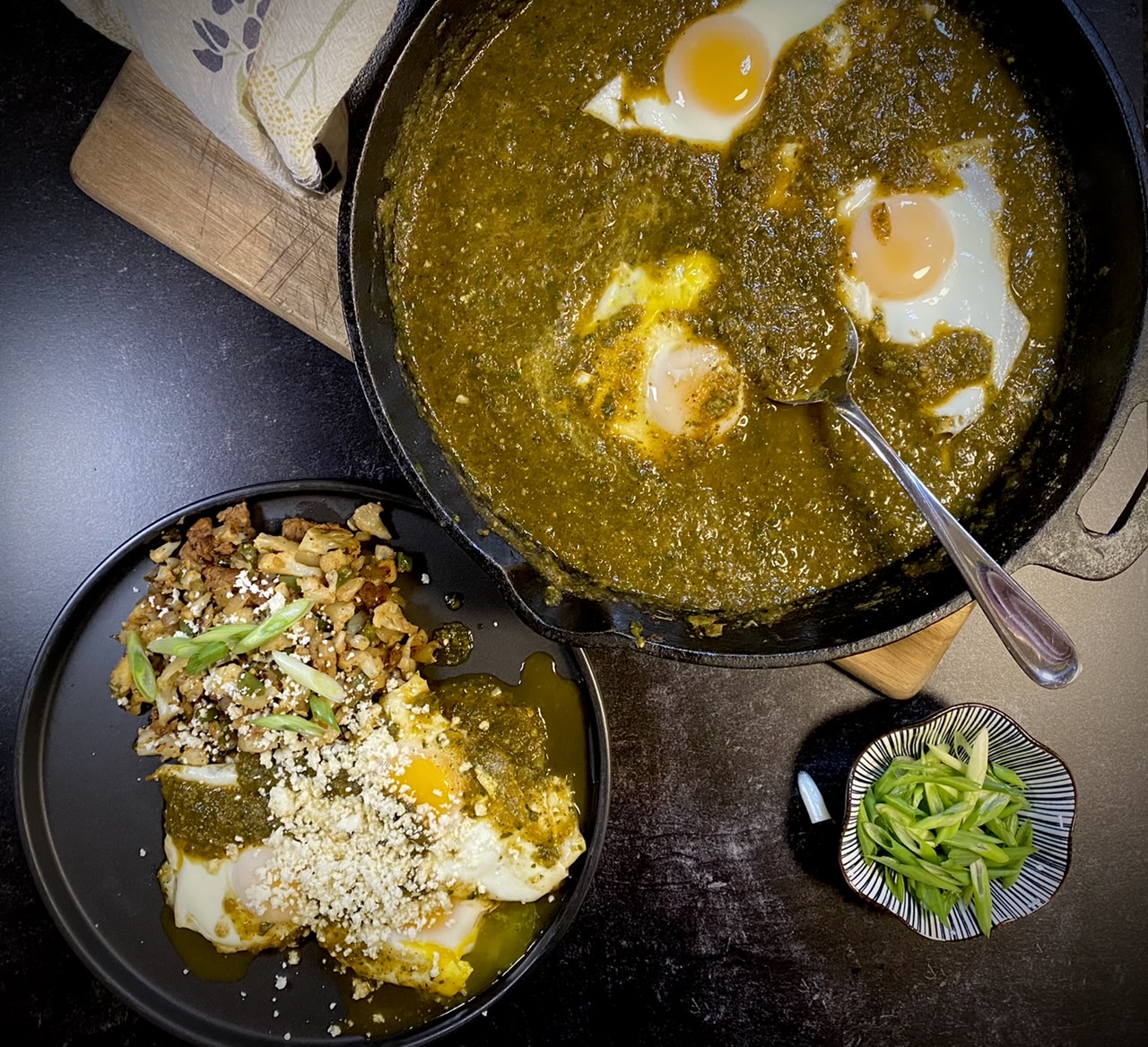 Green enchilada eggs and cauliflower hash on a black plate next to a cast iron pan of green enchilada eggs on top of a wooden cutting board
