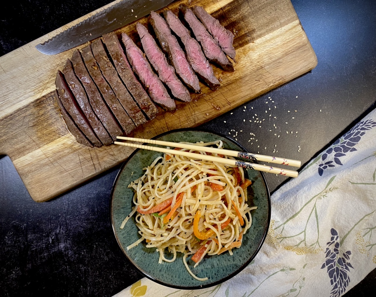 Flatiron steak sliced on a wooden cutting board next to a bowl of peanut noodles