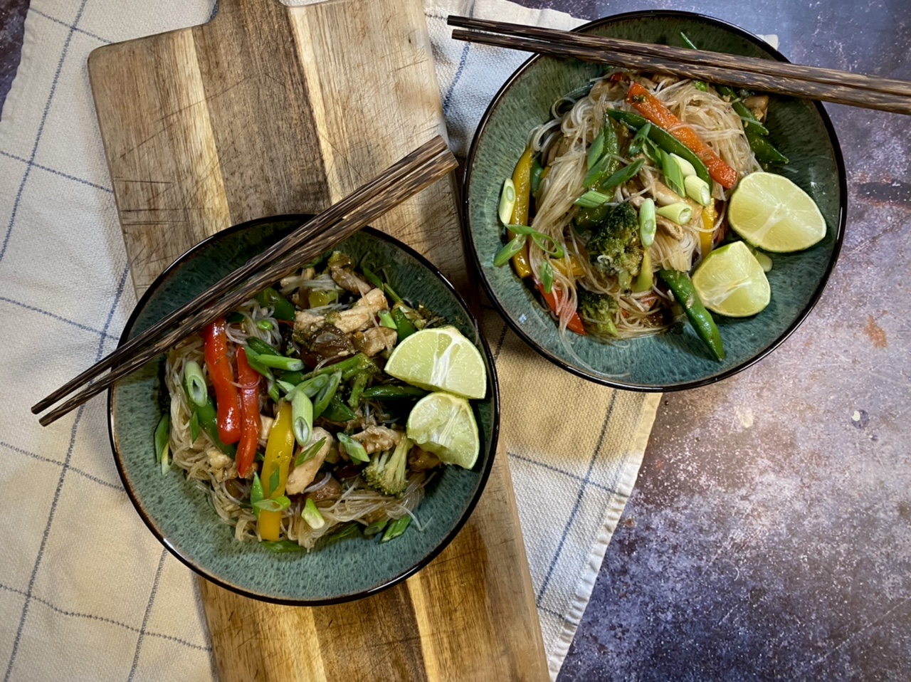Chicken stir fry in bowls with chopsticks on top of a wooden cutting board