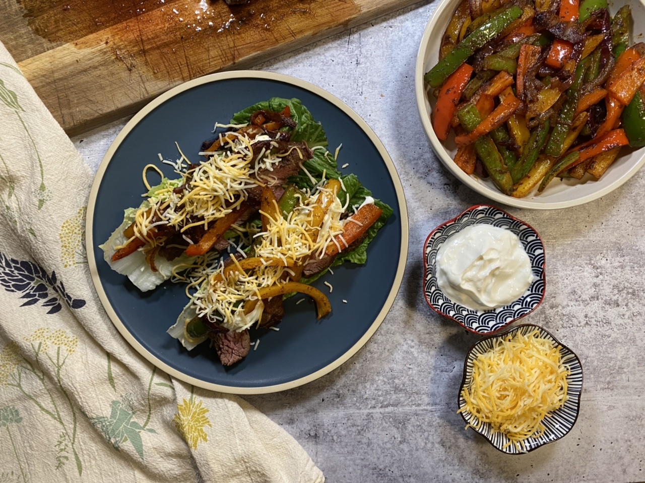 Romaine lettuce flank steak fajitas next to a cutting board of flank steak and a bowl of sautéed vegetables