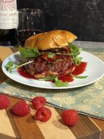 Venison burger with arugula and raspberry sauce on a white plate on top of a cutting board