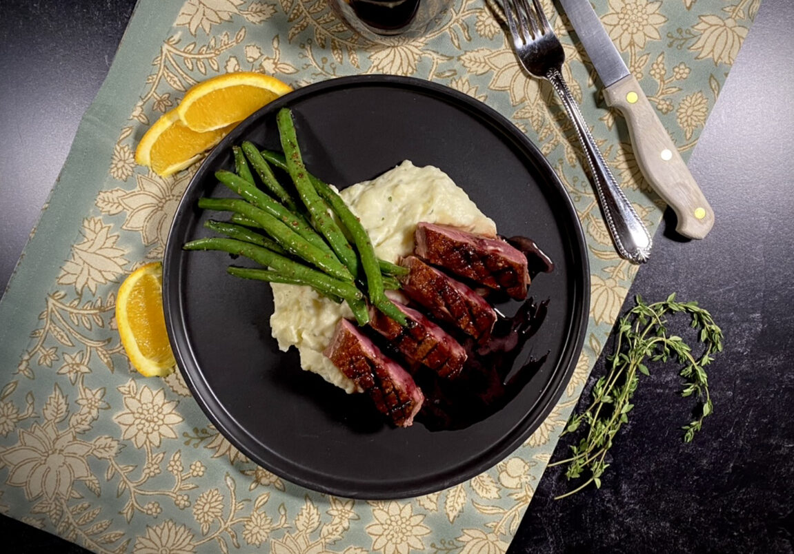 Sliced duck breast over mashed potatoes and green beans with pomegranate syrup on a black plate