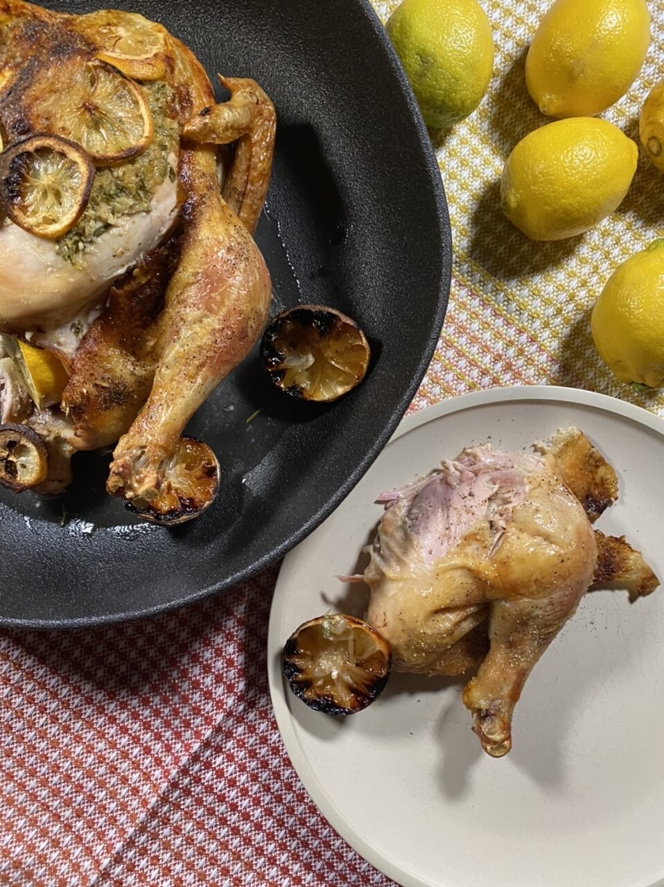 Whole roast chicken with lemons in a black bowl Next to a white plate with a chicken thigh