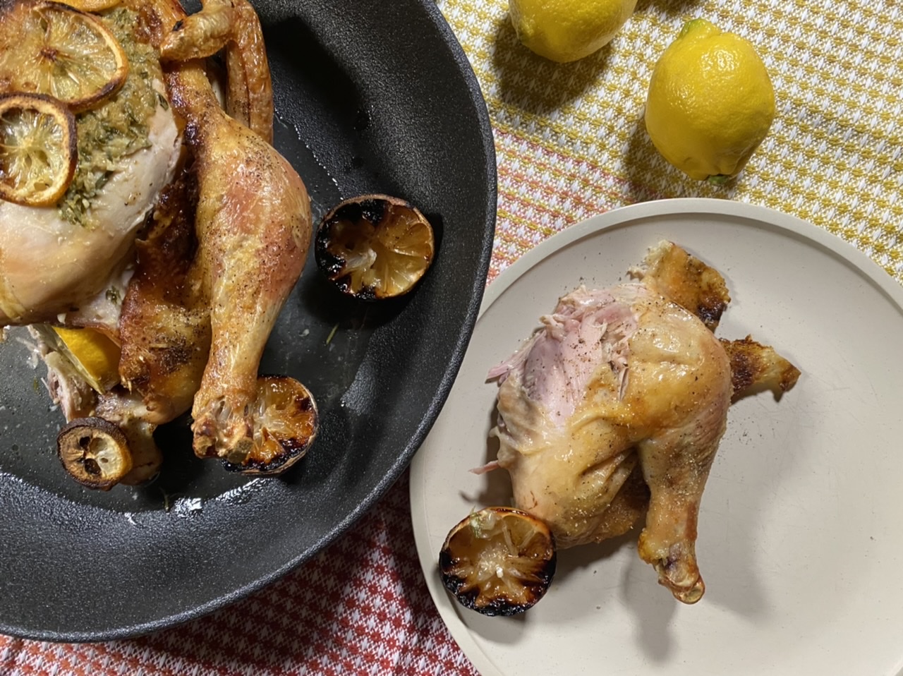 Whole roast chicken with lemons in a black bowl Next to a white plate with a chicken thigh