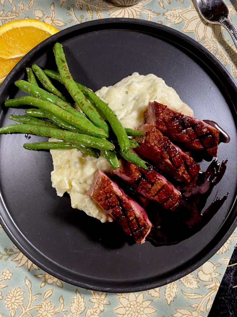 Sliced duck breast over mashed potatoes and green beans with pomegranate syrup on a black plate