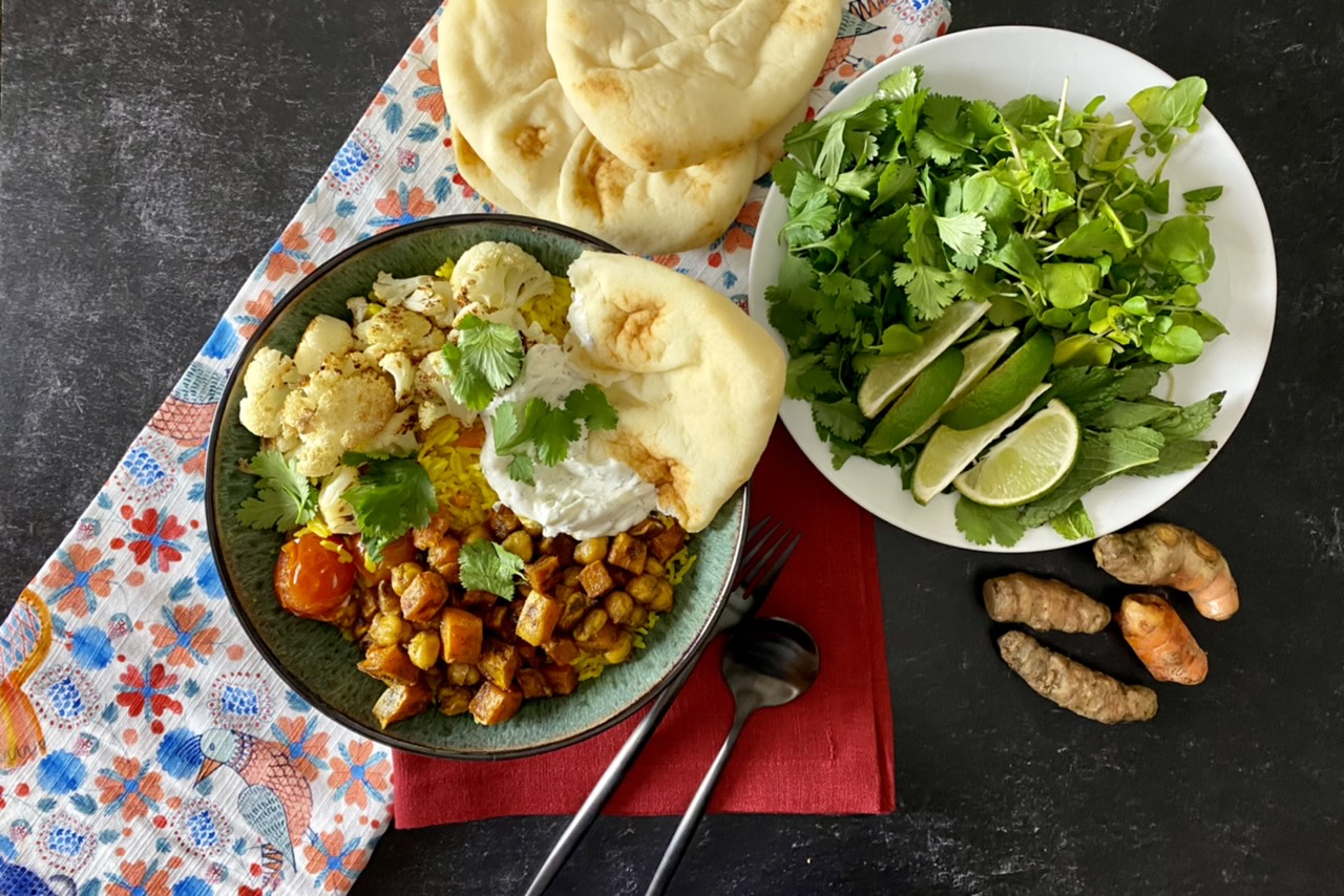 Turmeric rice with roasted cauliflower, chickpeas, and sweet potatoes, with burst cherries, tzatziki and naan in a green bowl