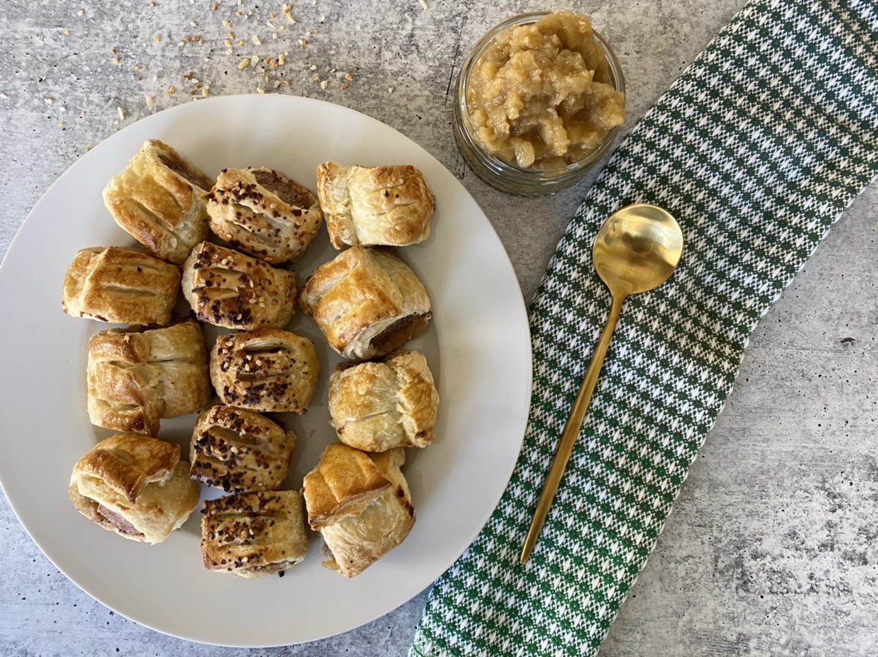 Sausage rolls on a white plate next to a green towel and a gold spoon with apple chutney