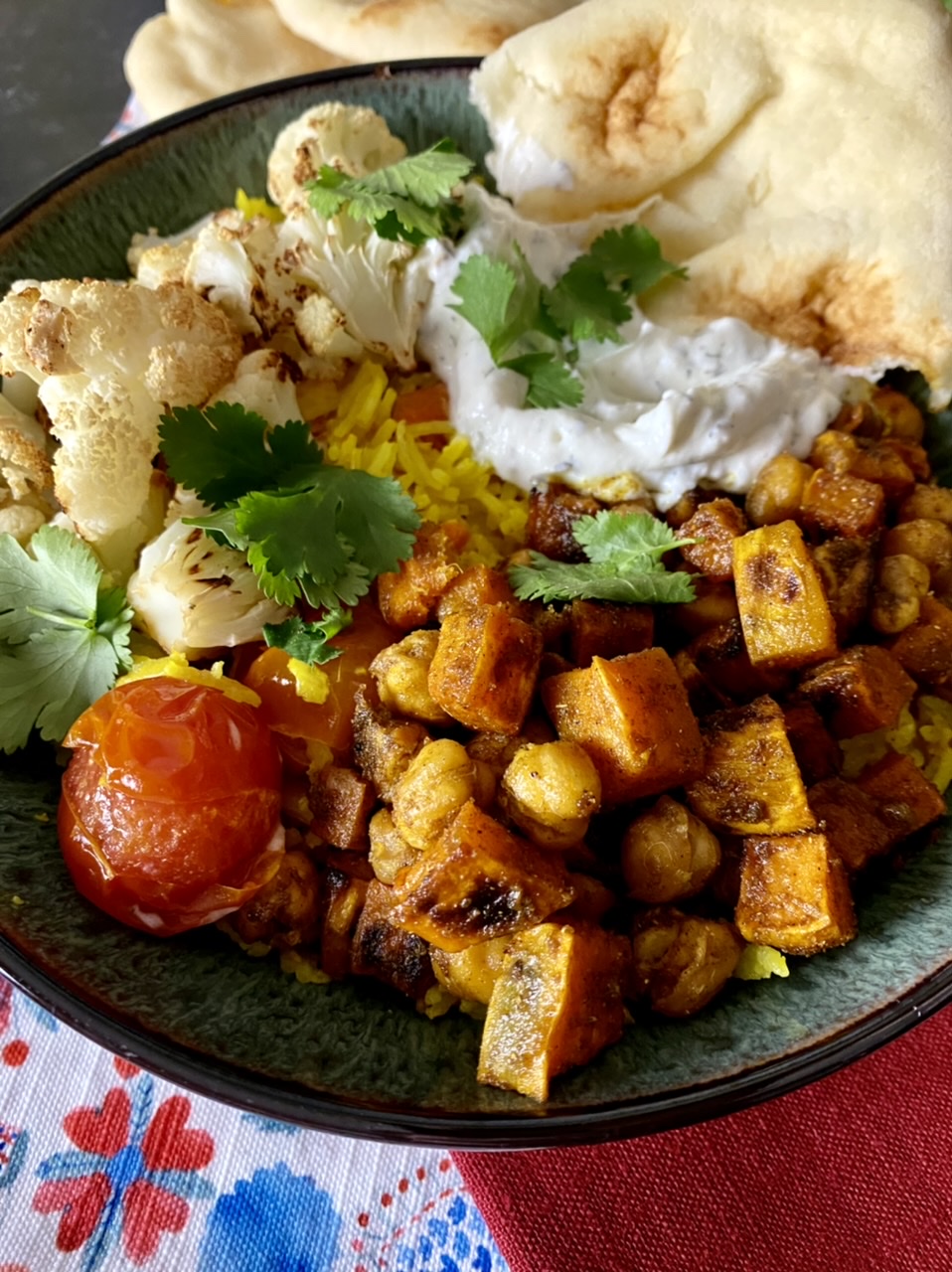 Turmeric rice with roasted cauliflower, chickpeas, and sweet potatoes, with burst cherries, tzatziki and naan in a green bowl