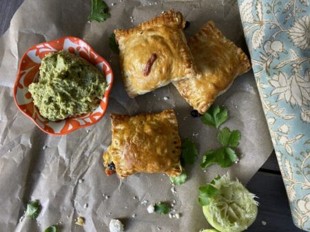 Mexican hand pies with black beans, corn, peppers and onions next to a bowl of avocado crema and squeezed limes and cilantro on top of parchment paper