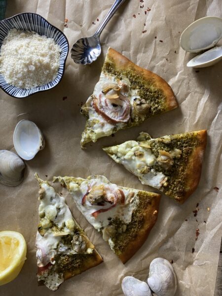 Pesto clam and prosciutto pizza on parchment paper next to a silver spoon and a bowl of Parmesan cheese and clam shells