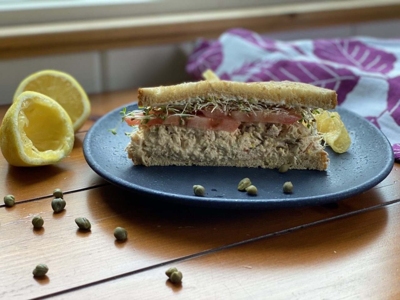 Tuna fish sandwich with browned butter aioli on a blue plate next to lemons and capers