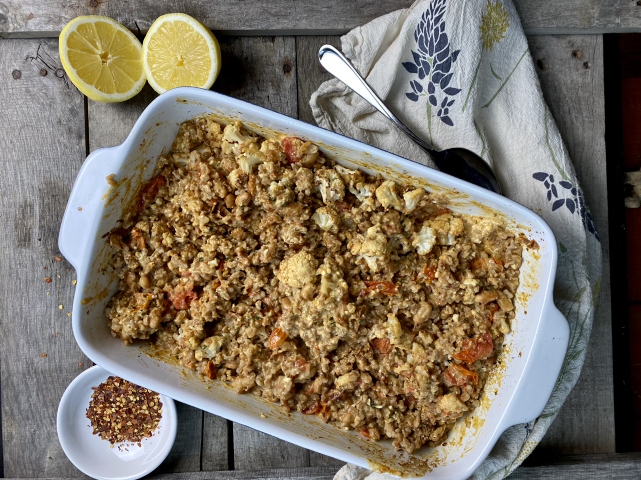 Baked feta and tomatoes with farro and roast cauliflower in a baking dish next to lemons and red pepper flakes