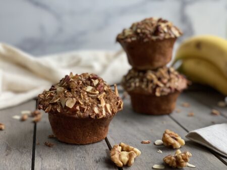 Three banana nut muffins with nut streusel next to walnuts, bananas, and a white towel