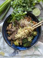 Korean Asian noodle bowl with broccoli, bean sprouts, and kimchi in a blue bowl next to a lime and cilantro