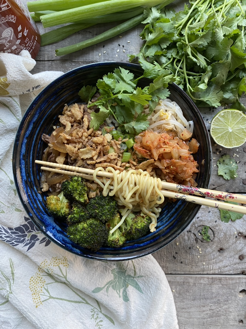 Korean Asian noodle bowl with broccoli, bean sprouts, and kimchi in a blue bowl next to a lime and cilantro