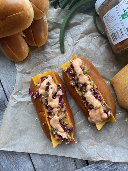 Korean hot dogs with kimchi slaw and gochujang mayonnaise on parchment paper next to scallions and hot dog buns and a mustard yellow napkin