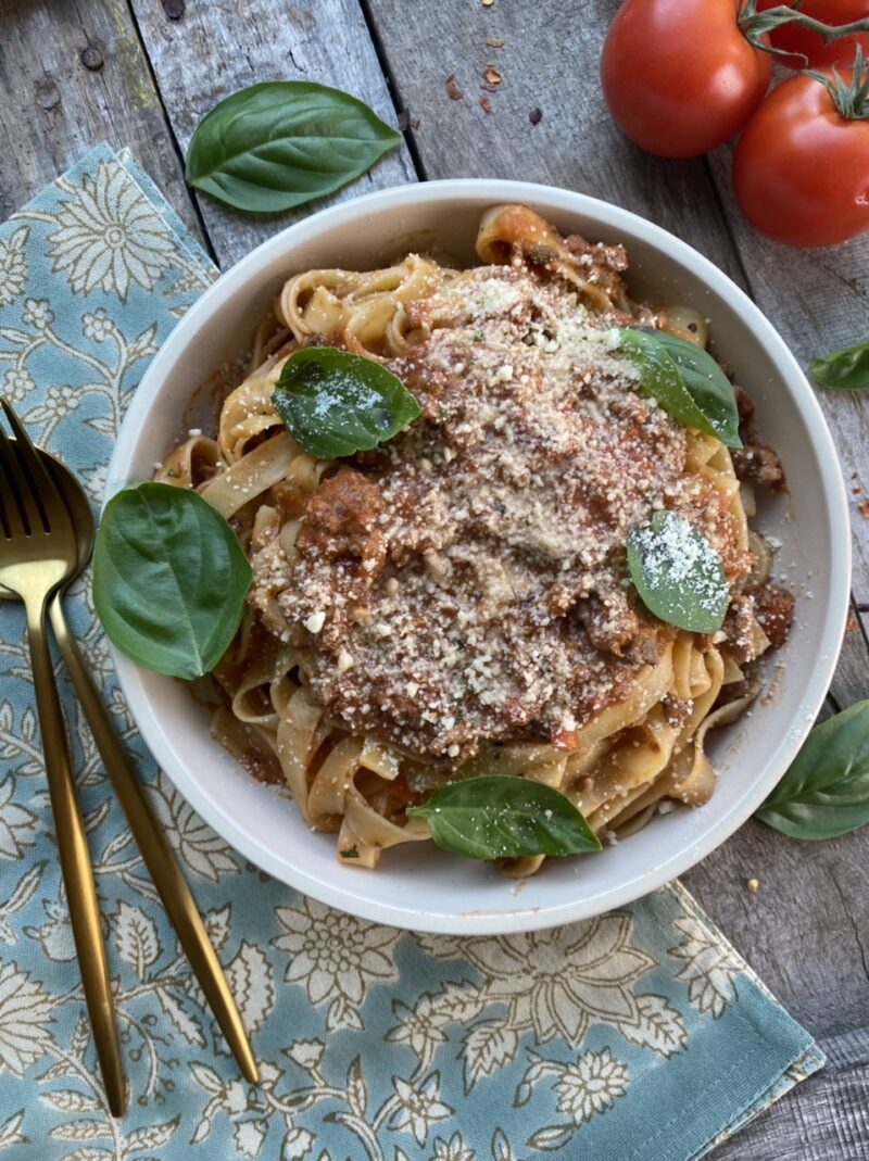 Homemade bolognese made from scratch with fresh basil in a white bowl next to gold silverware and tomatoes