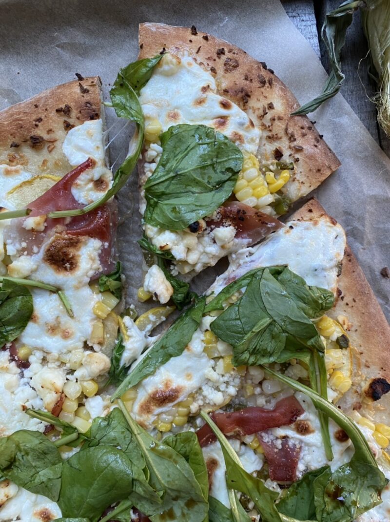 Summer corn, jalapeño, yellow squash, prosciutto, arugula, mozzarella, and feta pizza on parchment paper