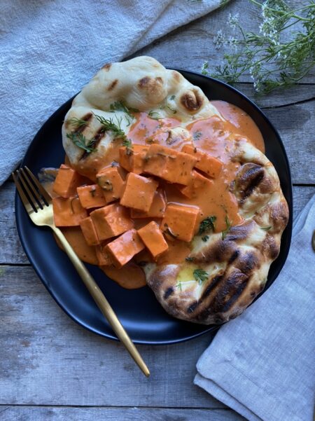Paneer tikka masala on top of grilled naan on a black plate with gold silverware